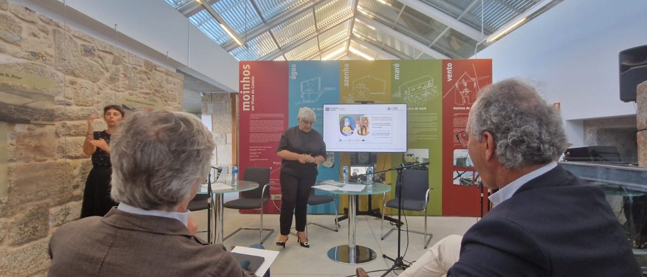 Imagem da interven&ccedil;&atilde;o de C&eacute;lia Sousa, de p&eacute;, virada para o p&uacute;blico, com o presidente da CIM Alto Alto Minho e o presidente da C&acirc;mara de Viana do Castelo sentados de costas, de frente para a palestrante